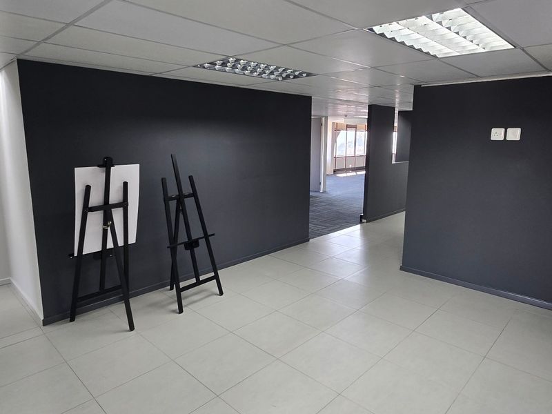 A spacious, open-plan commercial office area featuring neutral tiled flooring, dark accent walls, and a functional suspended ceiling with recessed lighting.
