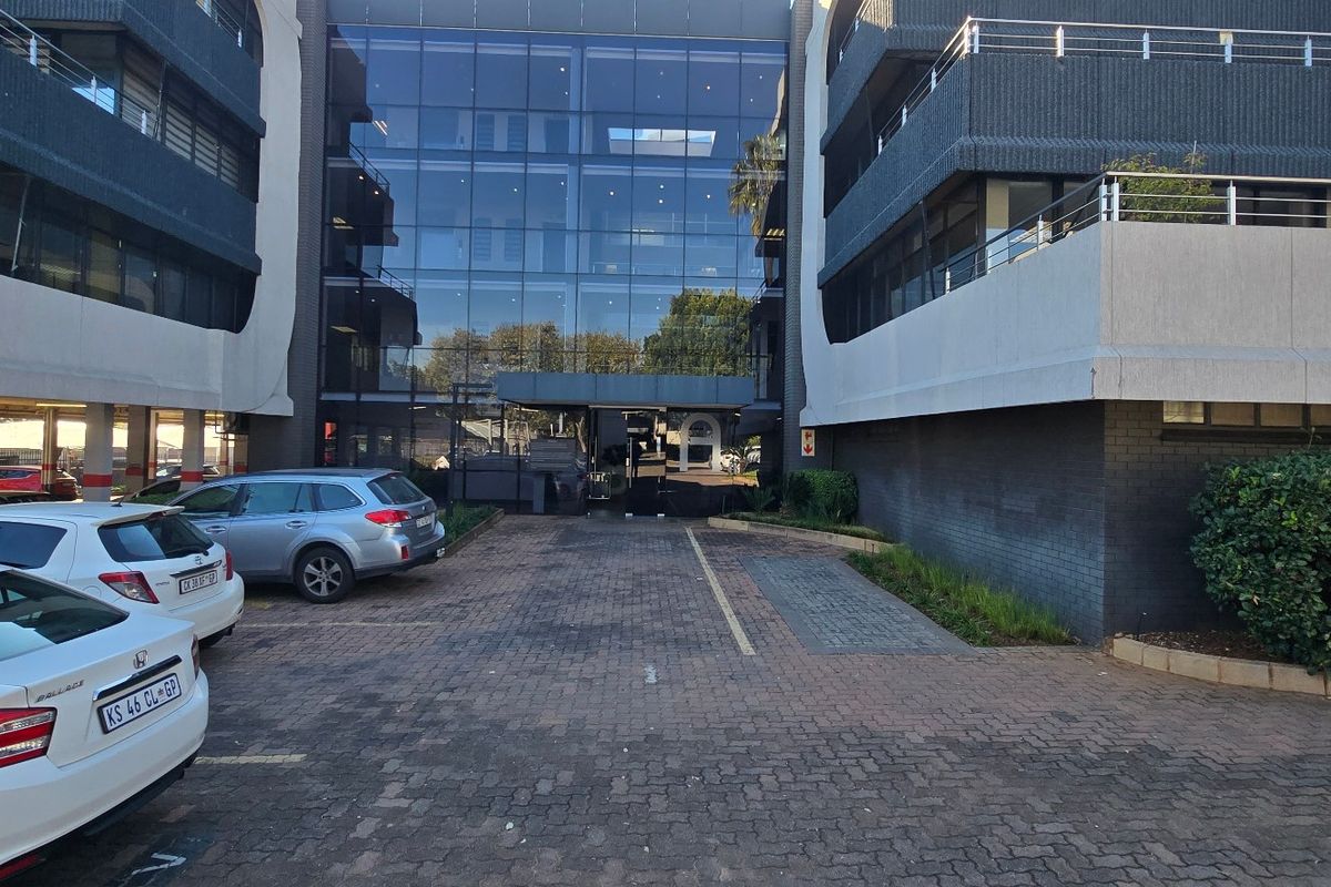 Front exterior view of a multi-story commercial office building featuring a glass facade, paved driveway, and covered parking facilities.