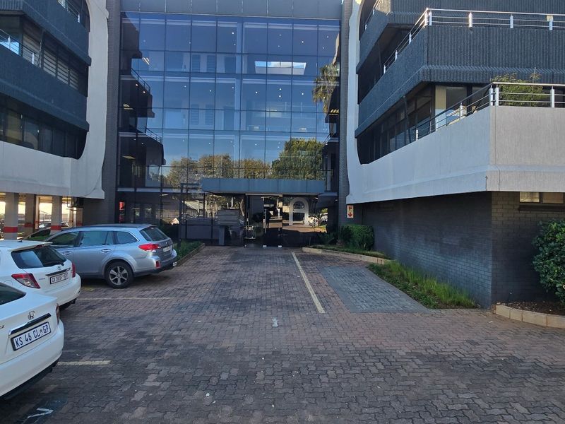 Front exterior view of a multi-story commercial office building featuring a glass facade, paved driveway, and covered parking facilities.