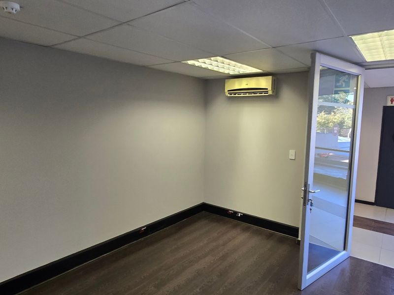 A clean, empty commercial office space featuring laminate flooring, a suspended ceiling with recessed lighting, and a wall-mounted air conditioning unit.