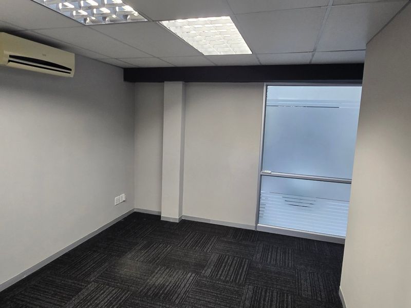 A clean, empty office space featuring modern carpet tiles, a suspended ceiling with recessed lighting, and a wall-mounted air conditioning unit.