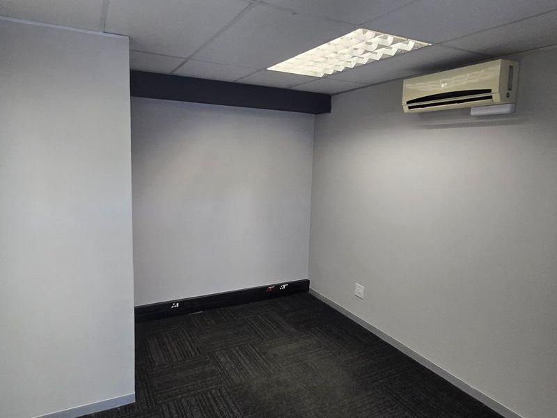 A clean, empty office space featuring carpet tiles, a suspended ceiling with recessed lighting, and a wall-mounted air conditioning unit.
