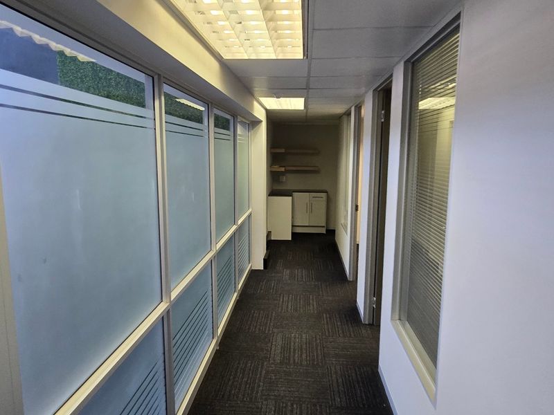 A clean commercial hallway featuring frosted glass partitions, recessed lighting, carpeted flooring, and a small built-in kitchenette or storage area at the end.