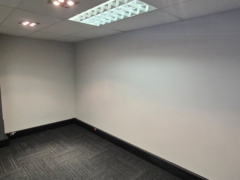 A clean, empty commercial office space featuring neutral walls, dark carpet tiles, and a suspended ceiling with integrated lighting.