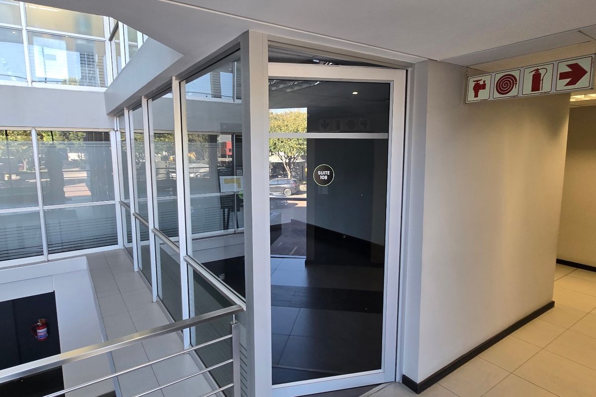 A modern commercial hallway featuring aluminium-framed glass partitioning, tiled flooring, and clear fire safety signage within a professional office building environment.