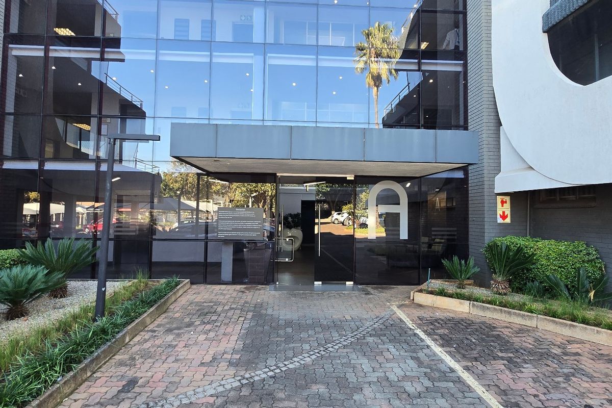 Modern commercial building entrance featuring a glass curtain wall facade, paved driveway, and well-maintained landscaped garden beds.