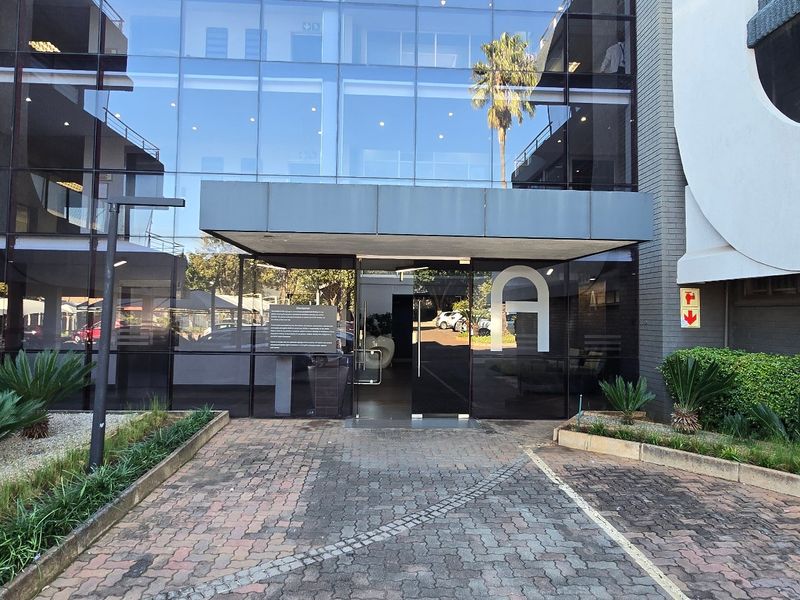 Modern commercial building entrance featuring a glass curtain wall facade, paved driveway, and well-maintained landscaped garden beds.