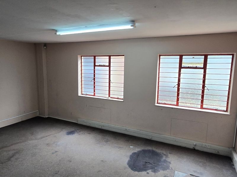 An empty industrial office space featuring fluorescent lighting, steel-framed windows with security bars, and surface-mounted electrical trunking.