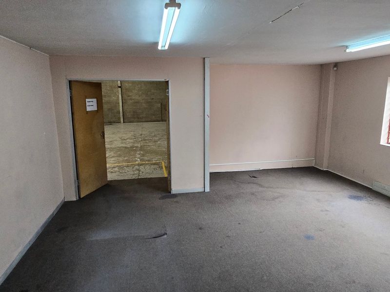 An empty office space featuring fluorescent lighting, security bars on the window, and worn carpet flooring, with direct access to a warehouse area.