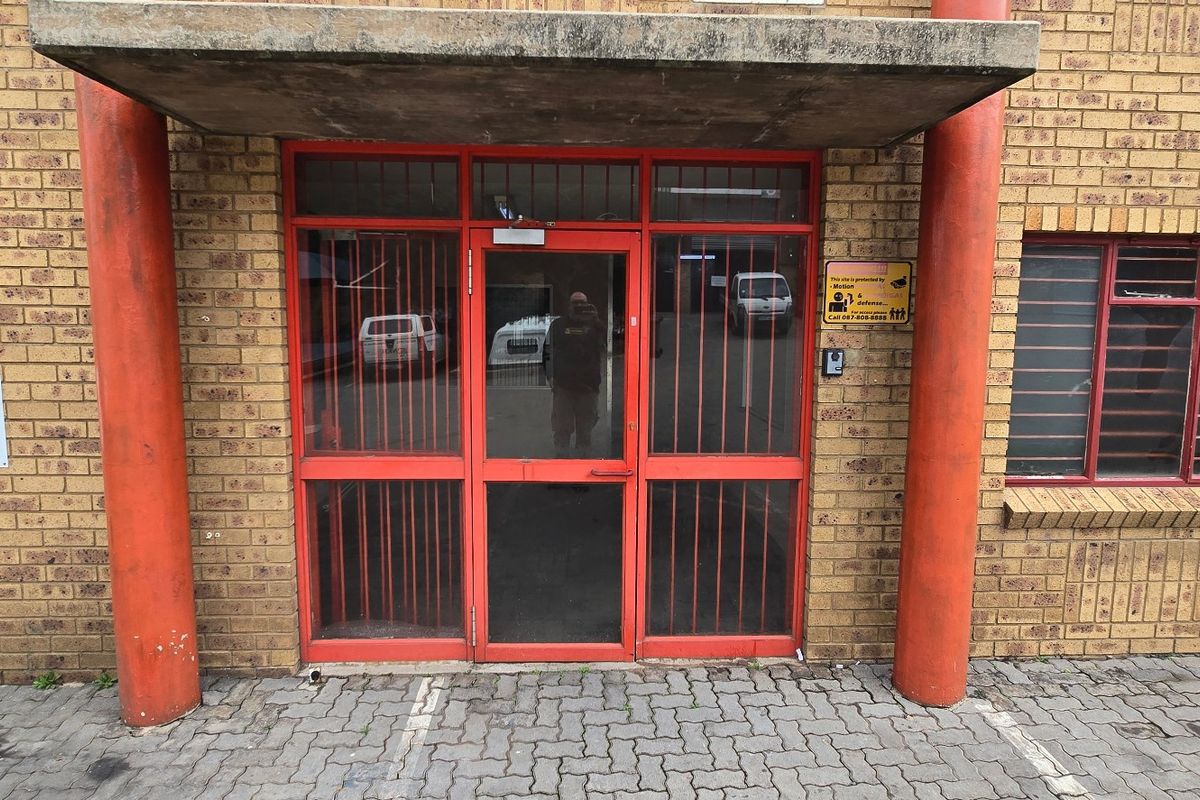 Front entrance of an industrial property featuring a brick facade, concrete canopy, and red metal security door and window frames.