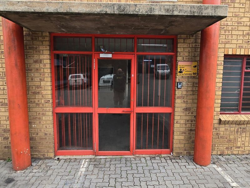 Front entrance of an industrial property featuring a brick facade, concrete canopy, and red metal security door and window frames.