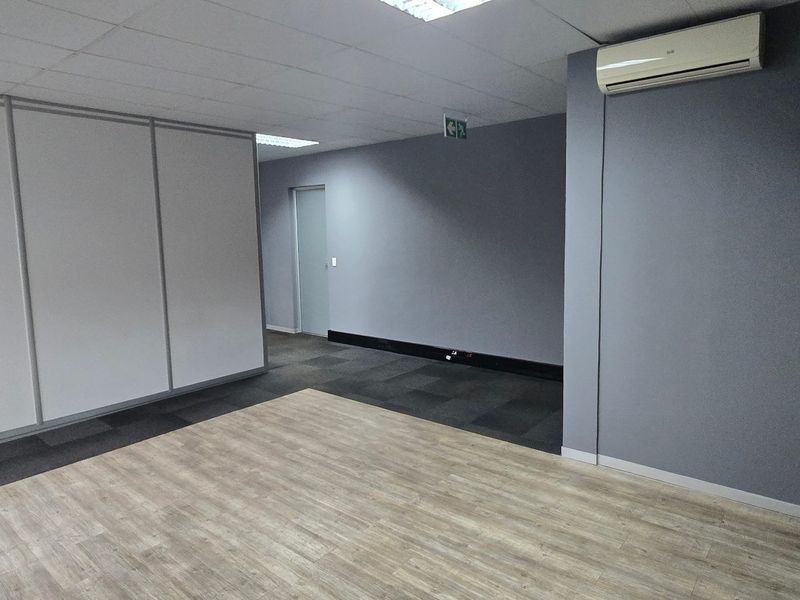 A clean, vacant commercial office space featuring a mix of laminate and carpet flooring, modular partition walls, and a wall-mounted air conditioning unit.