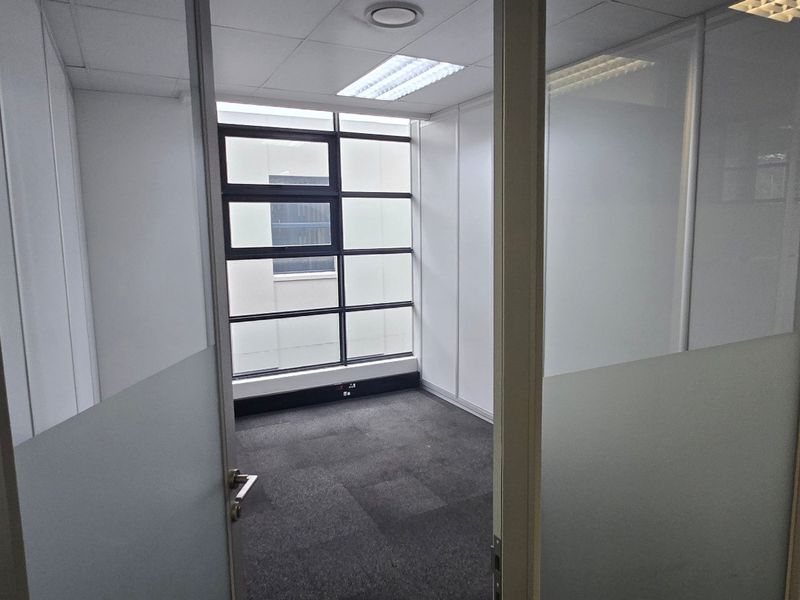 A vacant, modern office space featuring glass partition walls, carpeted flooring, and a large window providing natural light.