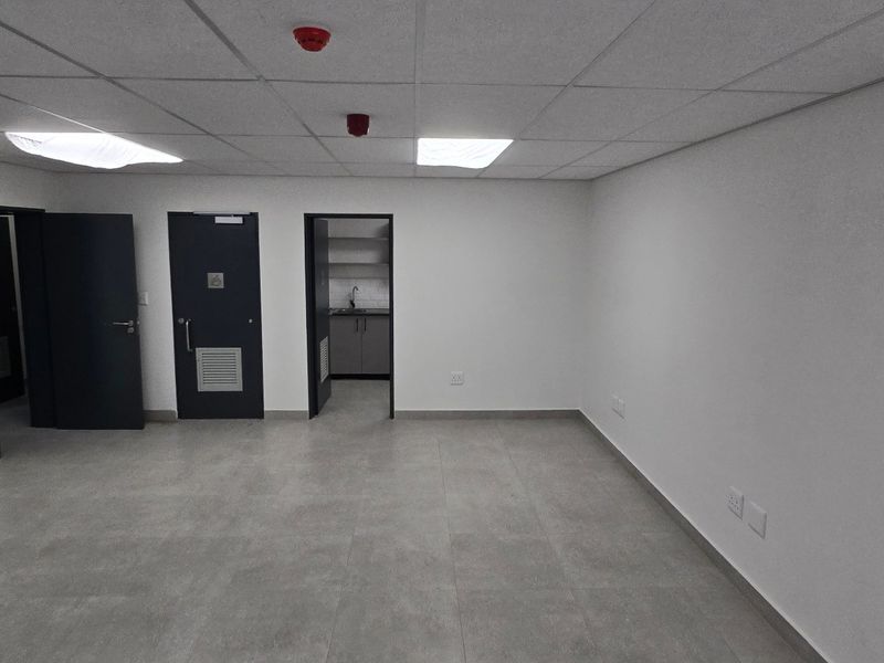 A clean, empty office space featuring neutral tiled flooring, a suspended ceiling with recessed lighting, and access to a small kitchenette area.