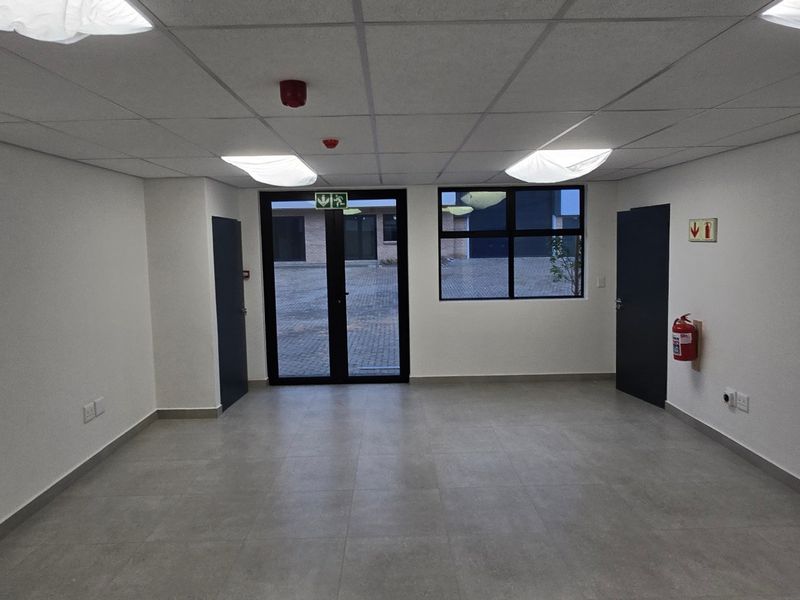 A clean, empty industrial office space featuring a suspended ceiling, recessed lighting, tiled floors, and direct access to an exterior paved area.