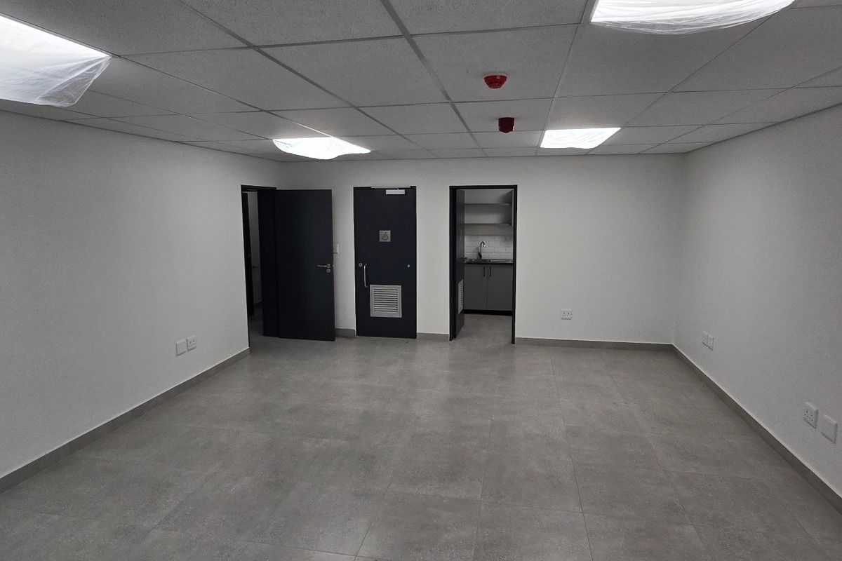 A clean, empty office space featuring modern tiled flooring, suspended ceilings with recessed lighting, and access to a small kitchenette area.