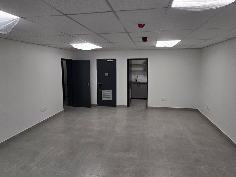 A clean, empty office space featuring modern tiled flooring, suspended ceilings with recessed lighting, and access to a small kitchenette area.