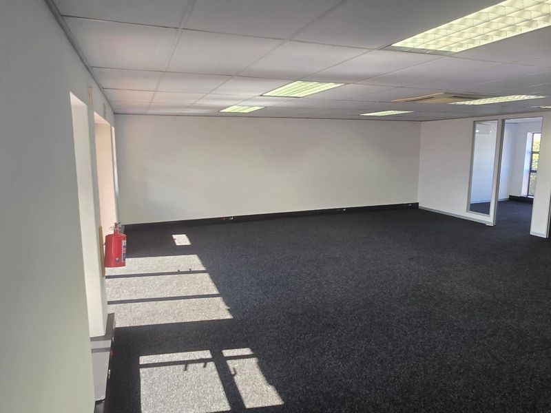 A spacious, vacant commercial office area featuring neutral walls, dark carpeting, suspended ceilings with recessed lighting, and glass-partitioned doorways.
