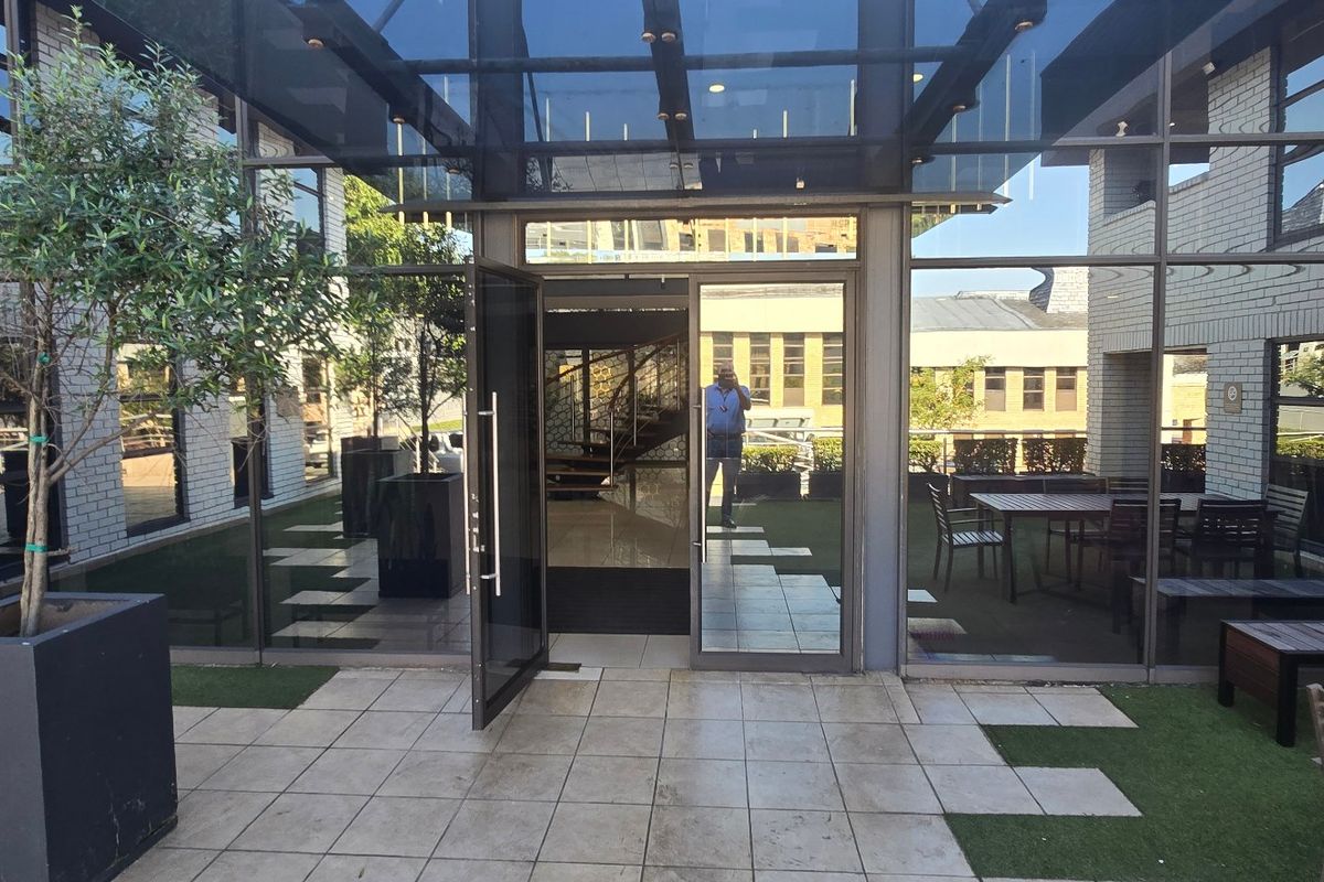 A modern commercial patio area featuring glass curtain walls, a glass canopy entrance, and a combination of tiled and artificial turf flooring.
