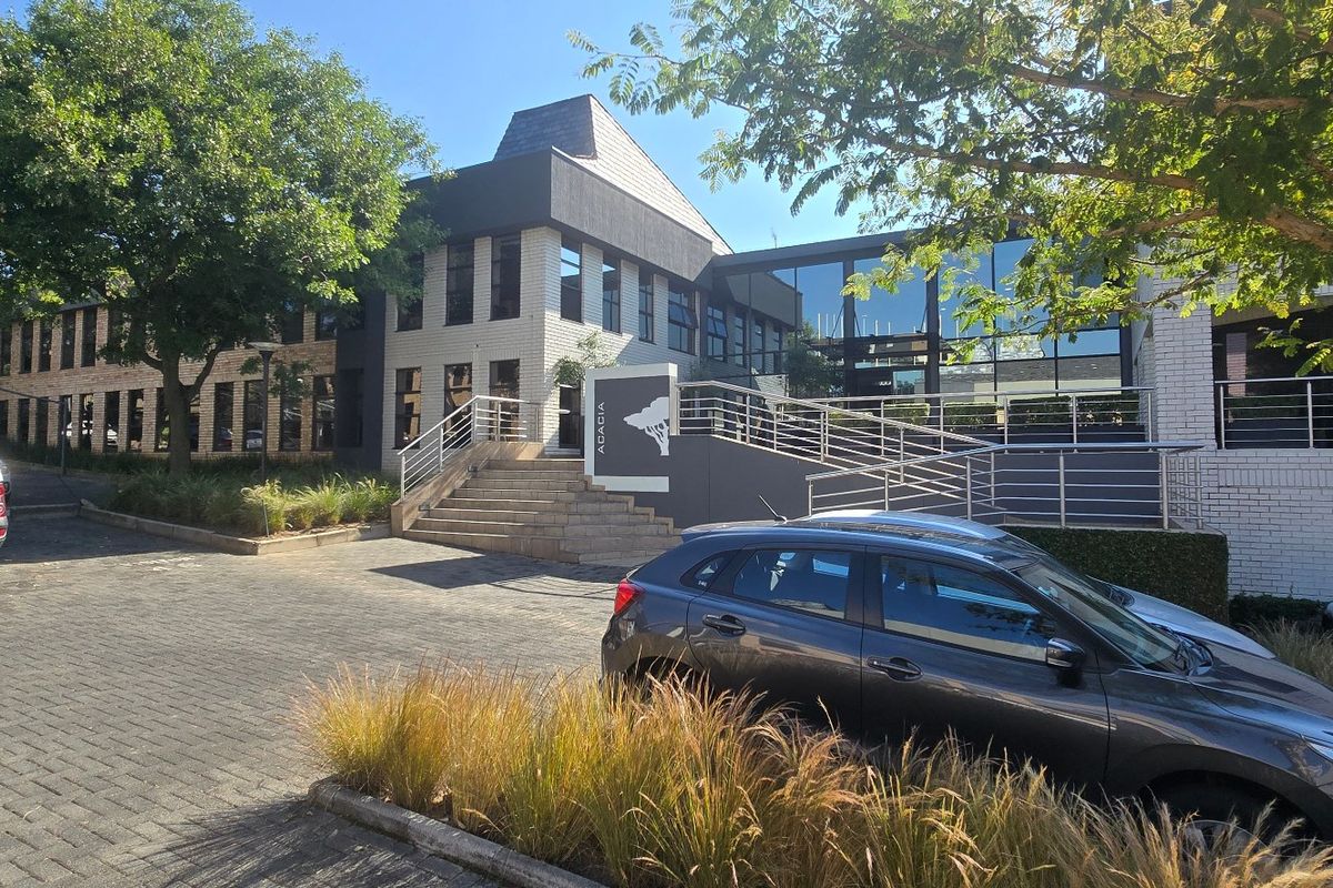 Front exterior view of a modern commercial office building featuring a paved parking area, concrete entrance stairs, and a glass facade.