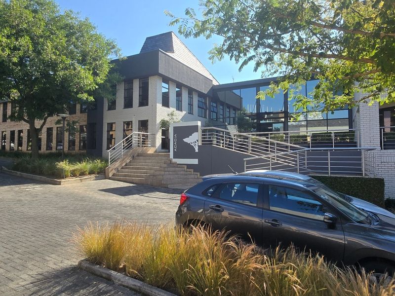 Front exterior view of a modern commercial office building featuring a paved parking area, concrete entrance stairs, and a glass facade.