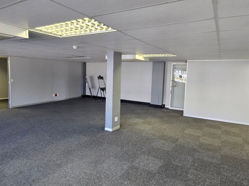 A spacious, open-plan commercial office area featuring neutral carpet tiles, a suspended ceiling with recessed lighting, and a central structural support column.