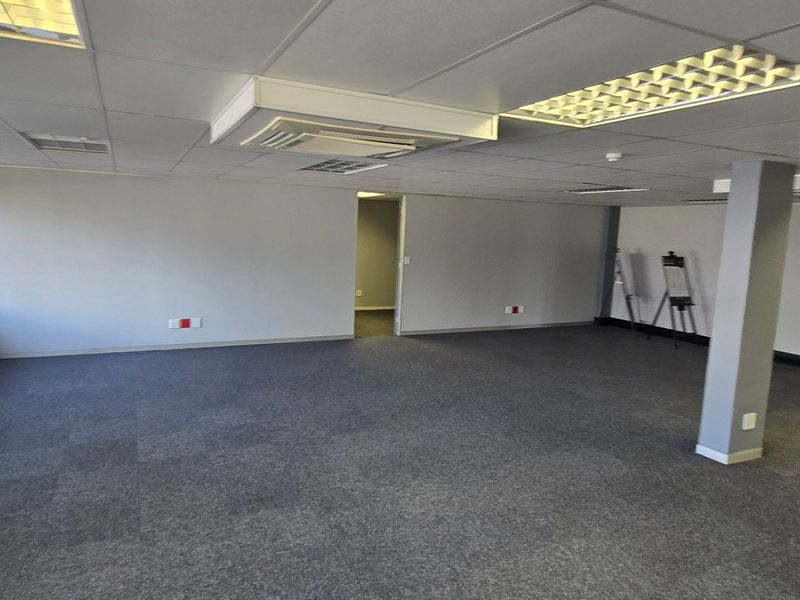 A spacious, open-plan commercial office area featuring carpet tiles, suspended ceilings with recessed lighting, and large windows providing natural light.