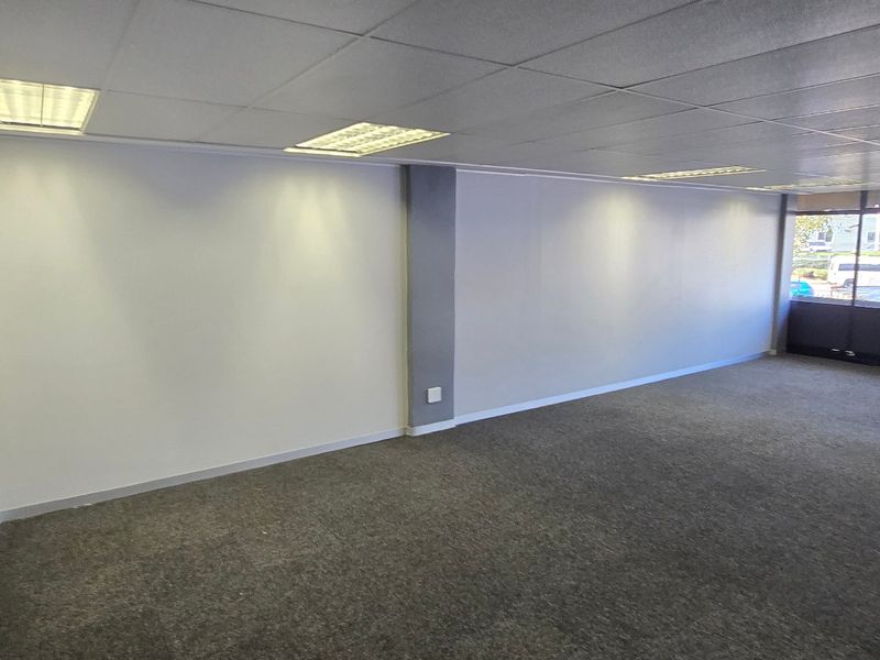 A spacious, empty commercial office area featuring neutral walls, carpeted flooring, and large windows providing ample natural light.