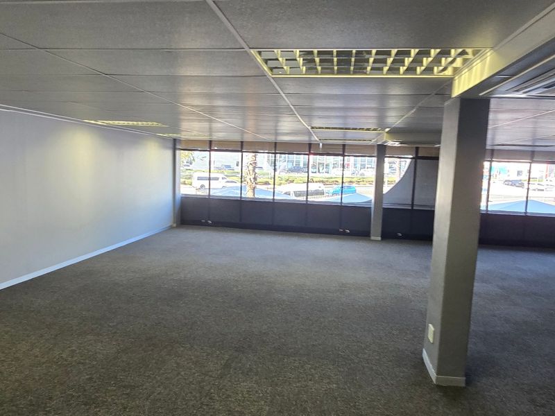 Spacious, open-plan commercial office area featuring carpeted flooring, suspended ceilings with recessed lighting, and large windows providing ample natural light.