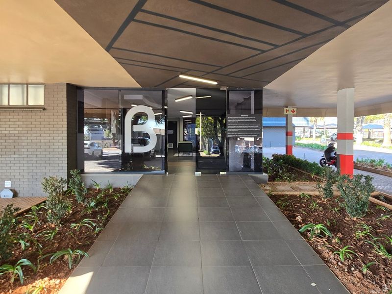 A modern commercial building entrance featuring a covered walkway, glass doors, and landscaped garden beds leading to the main reception area.