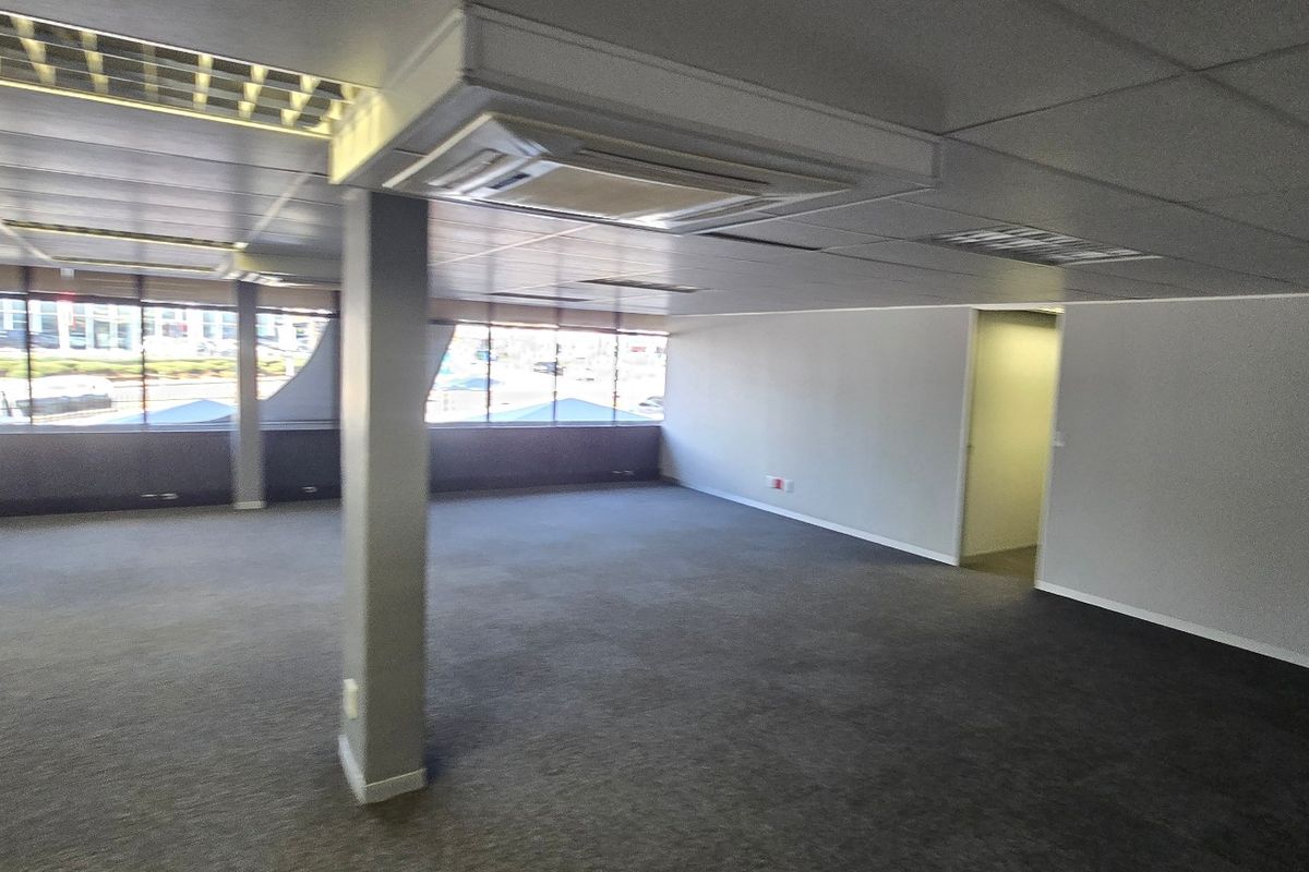 A spacious, open-plan commercial office area featuring large windows, carpeted flooring, a suspended ceiling with recessed lighting, and integrated central air conditioning.