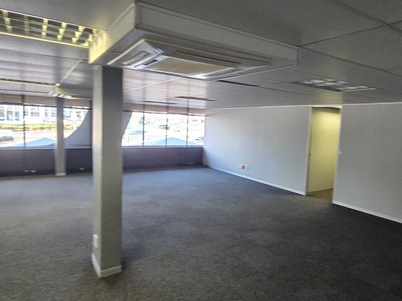 A spacious, open-plan commercial office area featuring large windows, carpeted flooring, a suspended ceiling with recessed lighting, and integrated central air conditioning.