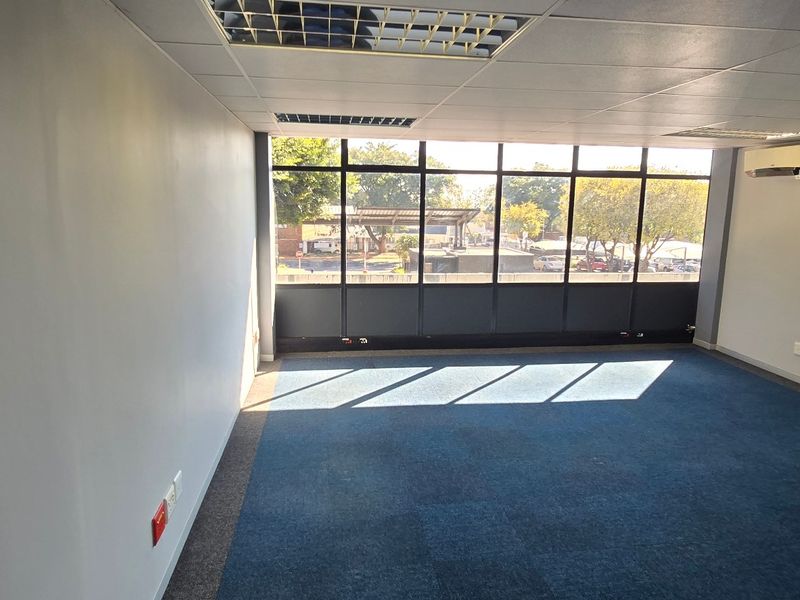 A bright, empty commercial office space featuring large windows, blue carpeting, a suspended ceiling with recessed lighting, and a wall-mounted air conditioning unit.