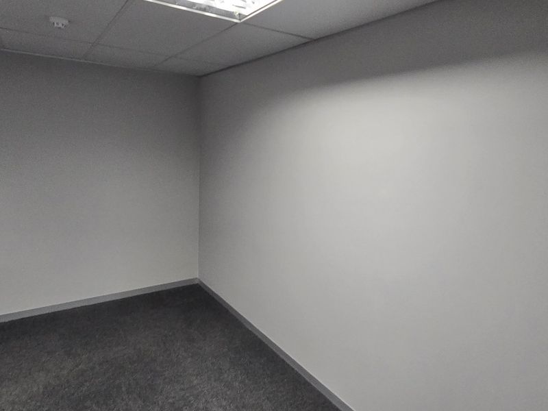 A clean, empty office space featuring neutral walls, carpeted flooring, and a suspended ceiling with recessed lighting.