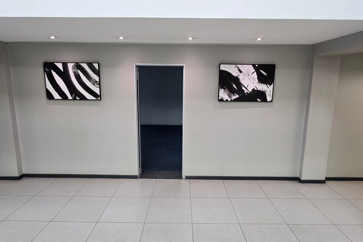 A clean, modern hallway featuring neutral tiled flooring, recessed downlighting, and a central doorway leading to an adjacent office space.