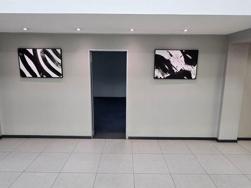 A clean, modern hallway featuring neutral tiled flooring, recessed downlighting, and a central doorway leading to an adjacent office space.