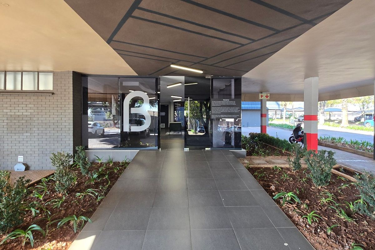 Modern commercial building entrance featuring a covered walkway, glass doors, and well-maintained landscaped garden beds.