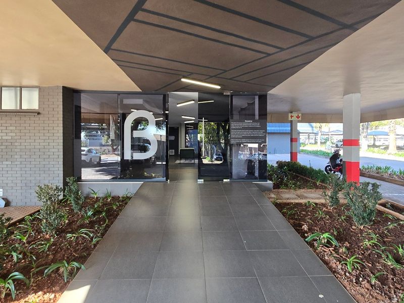 Modern commercial building entrance featuring a covered walkway, glass doors, and well-maintained landscaped garden beds.