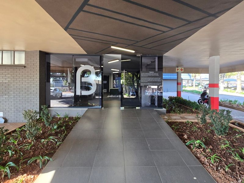 A modern commercial building entrance featuring a covered walkway, glass doors, tiled flooring, and well-maintained landscaped garden beds.
