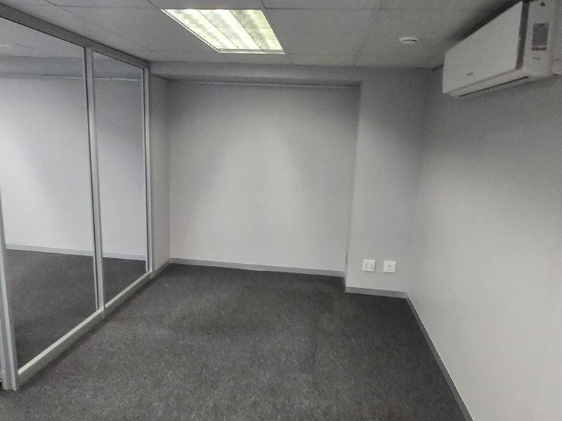 A clean, empty office space featuring glass partitioning, carpeted flooring, recessed ceiling lighting, and a wall-mounted air conditioning unit.