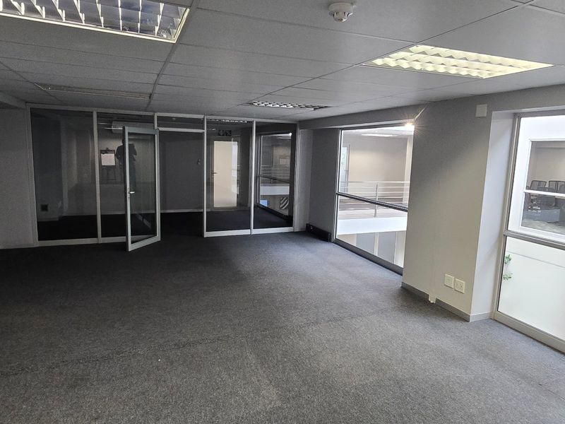 A bright, open-plan office space featuring glass partitions, carpeted flooring, recessed lighting, and a decorative artificial green wall.