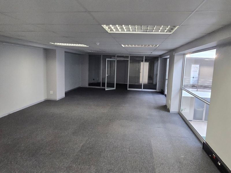 A spacious, empty commercial office area featuring carpeted flooring, suspended ceilings with recessed lighting, and glass partitions, offering a clean and professional workspace.
