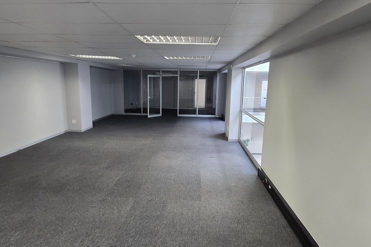 A spacious, empty commercial office area featuring a suspended ceiling, recessed lighting, carpeted flooring, and glass partitions, offering a clean and professional workspace.
