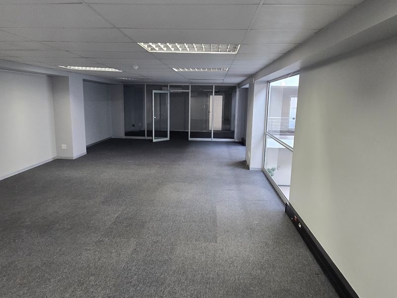 A spacious, empty commercial office area featuring a suspended ceiling, recessed lighting, carpeted flooring, and glass partitions, offering a clean and professional workspace.