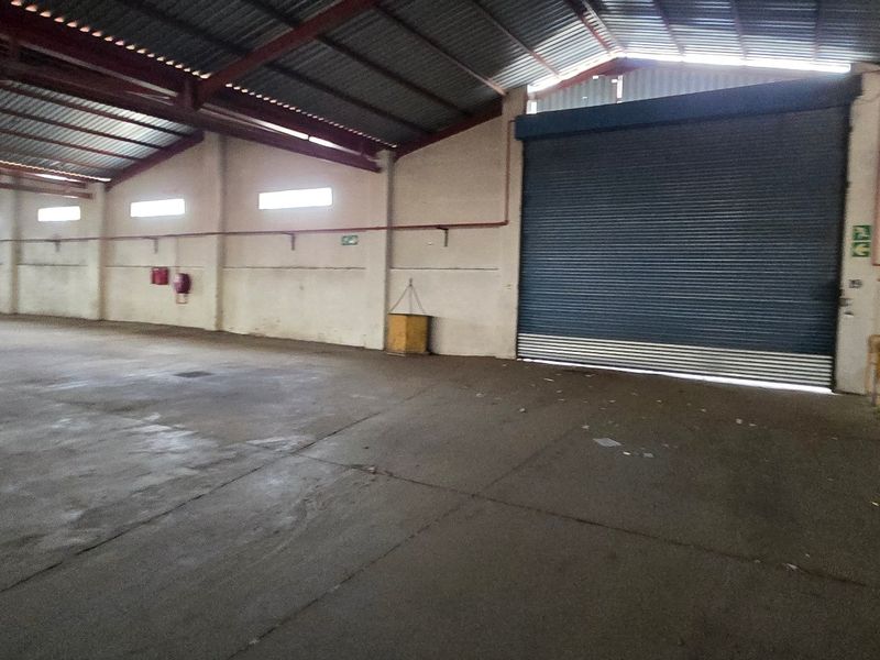 Spacious industrial warehouse unit featuring high ceilings with exposed steel trusses, concrete flooring, and a large roller shutter door for easy access.