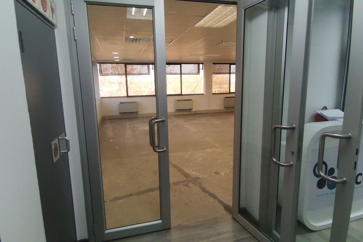 An empty commercial office space featuring large windows, suspended ceilings, and wall-mounted HVAC units, accessible via double glass doors.