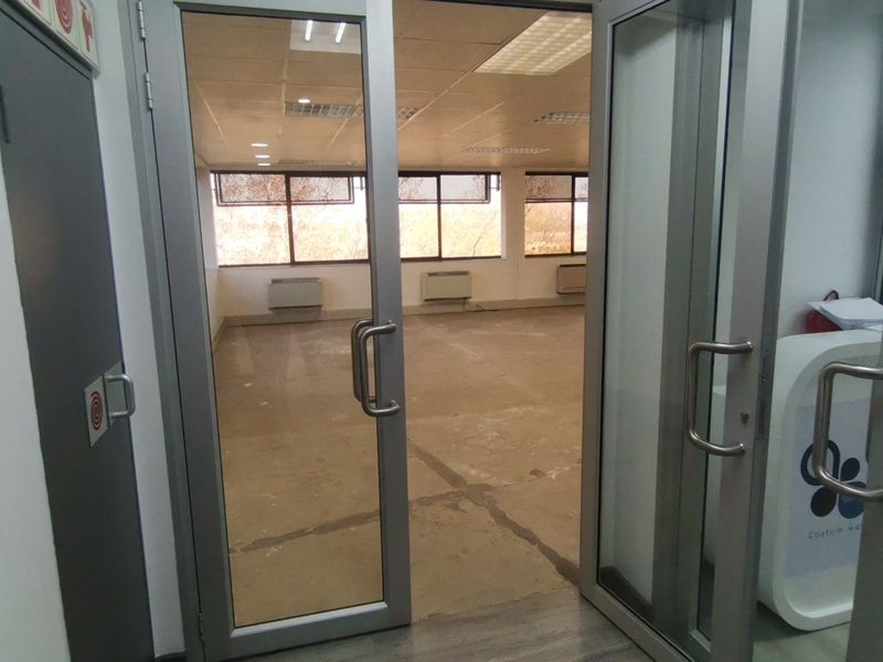 An empty commercial office space featuring large windows, suspended ceilings, and wall-mounted HVAC units, accessible via double glass doors.