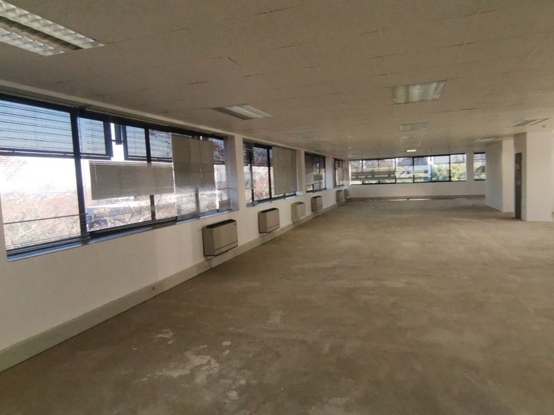 A large, open-plan commercial office space featuring expansive windows, wall-mounted air conditioning units, and a suspended ceiling, currently in a vacant, shell-like condition.
