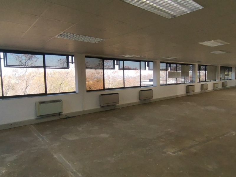 A large, vacant open-plan office space featuring expansive windows, wall-mounted air conditioning units, and a suspended ceiling with recessed lighting.