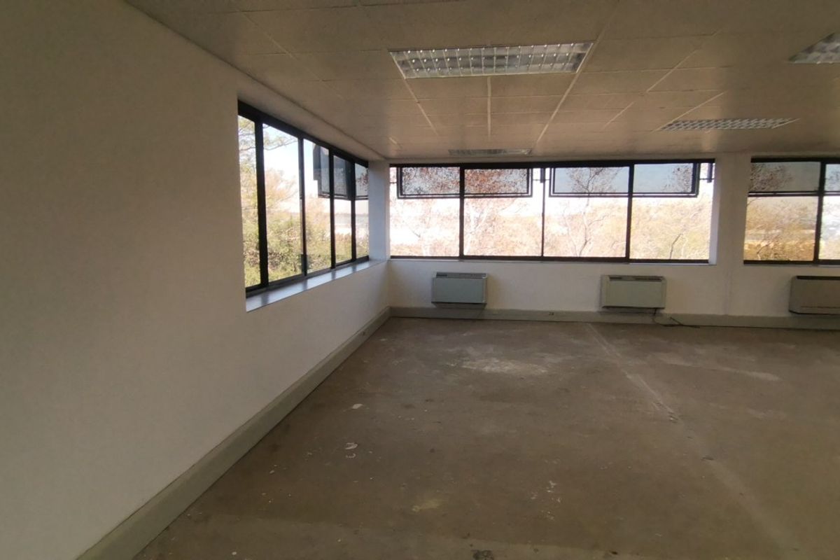An empty, open-plan commercial office space featuring large windows, wall-mounted HVAC units, and a suspended ceiling with recessed lighting.